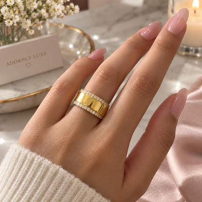 A wide gold ring with two rows of small clear stones, show it on a model engagement finger The scene includes soft pink fabric, white baby's breath flowers, and a brand card in display. 
