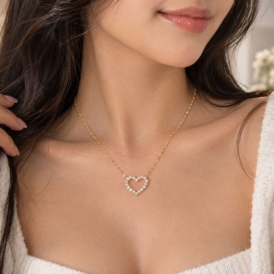 A close-up lifestyle shot of a model wearing a delicate gold heart-shaped pendant necklace adorned with small clear stones, paired with a white knit top.