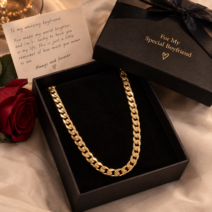 A gold chain necklace displayed in an open black presentation box with a black ribbon and a handwritten note beside it, styled on a soft beige fabric with a red rose.

