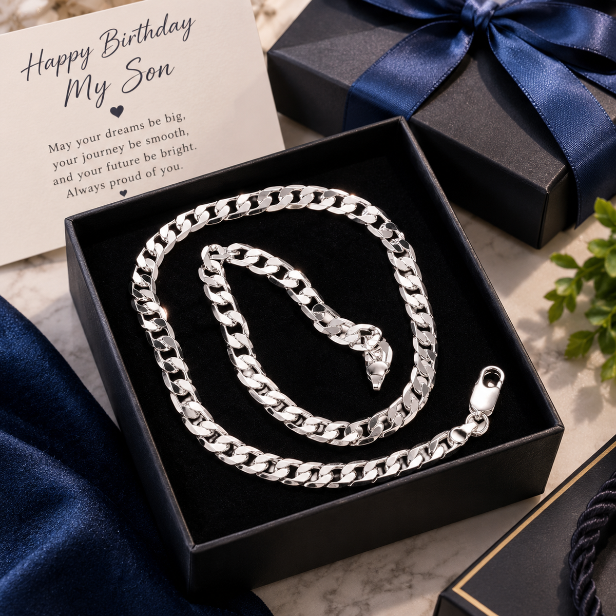 A silver Cuban link chain necklace presented in an open black gift box with a velvet insert, alongside a printed birthday card that reads 'Happy Birthday My Son' and additional black gift boxes with blue ribbons on a marble surface.
