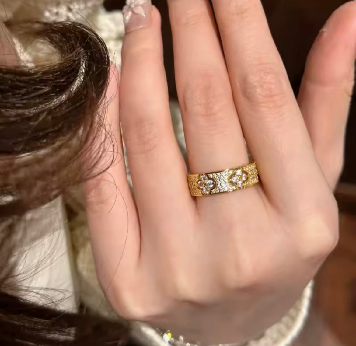 A close-up of a hand wearing a gold band ring with intricate floral or vine engravings and small clear stones, photographed indoors against a softly blurred dark background.