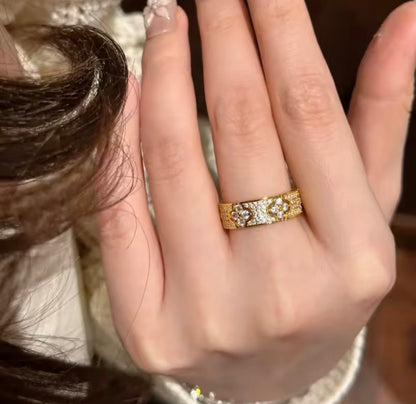 A close-up of a hand wearing a gold band ring with intricate floral or vine engravings and small clear stones, photographed indoors against a softly blurred dark background.