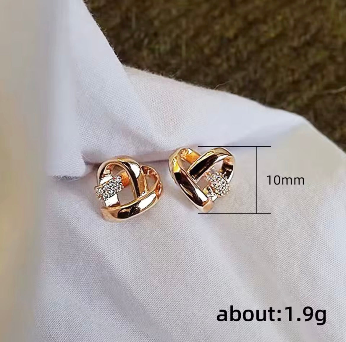 A pair of rose gold heart-shaped stud earrings with small clear crystal accents, displayed on a white fabric background with a dimension line indicating 10mm size and text reading about: 1.9g.