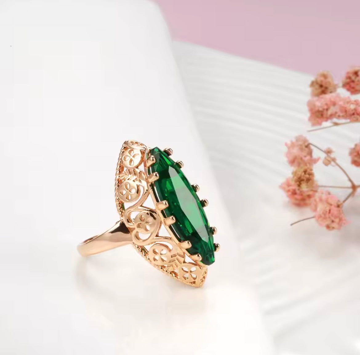 A close-up of a hand wearing a gold ring with a marquise-cut green emerald gemstone, set against a soft-focus background of flowers and a marble surface.