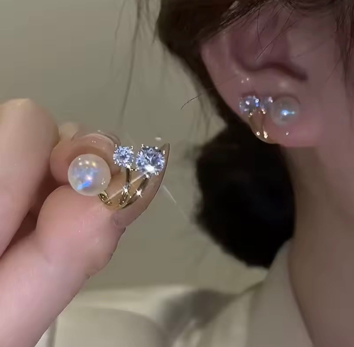A close-up lifestyle shot of a person wearing gold crescent-shaped ear cuffs with white iridescent pearls and clear rhinestones, with a hand holding the unstyled cuff next to their ear.