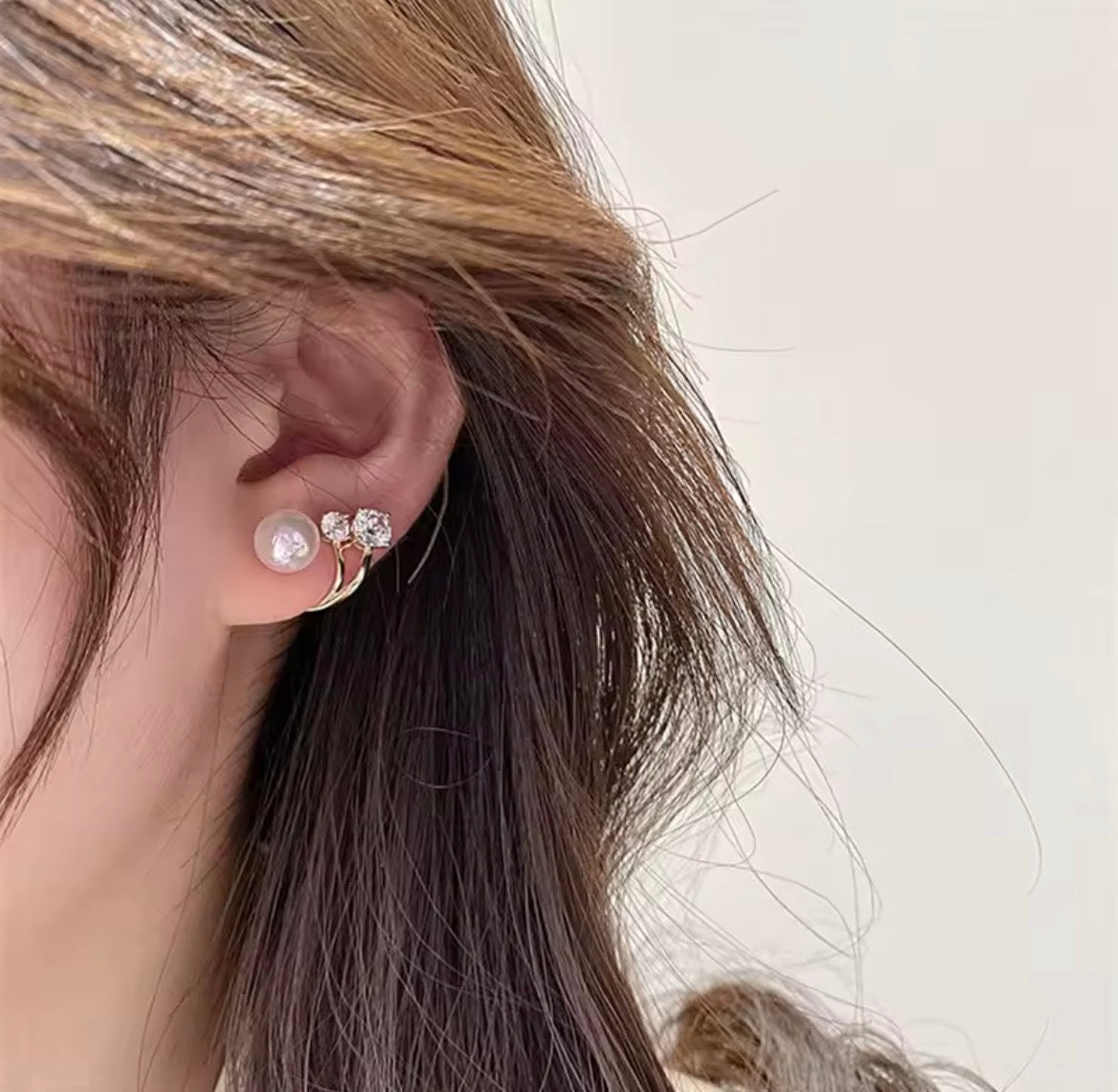 A close-up of a person's ear wearing a small earring set with a white pearl and three clear rhinestones, with brown hair framing the side of the face against a light neutral background.