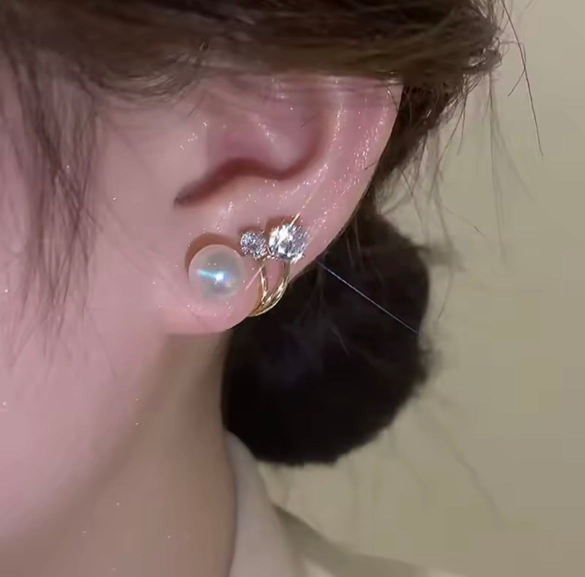 Close-up of a person's ear wearing a gold ear cuff with a large iridescent pearl and small clear crystal charms, against a plain beige background.