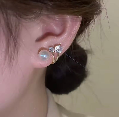 Close-up of a person's ear wearing a gold ear cuff with a large iridescent pearl and small clear crystal charms, against a plain beige background.