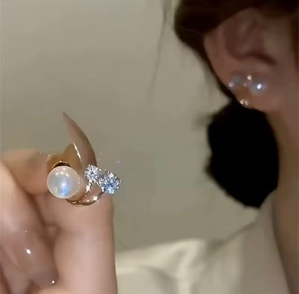 A close-up of a hand holding a gold ring with a large white pearl and a small cluster of clear stones, with part of a person's ear and face visible in the background.

