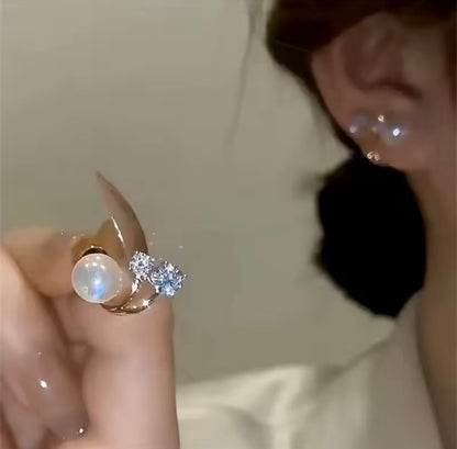A close-up of a hand holding a gold ring with a large white pearl and a small cluster of clear stones, with part of a person's ear and face visible in the background.

