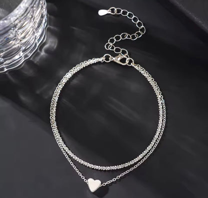 A silver double-strand chain anklet with a small heart charm, displayed on a dark reflective surface.