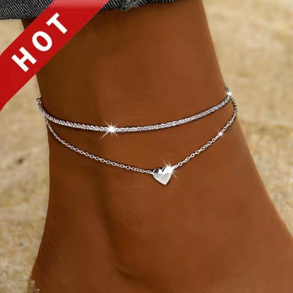 A close-up of a person's ankle wearing two delicate silver chain anklets, one with a small heart charm and one with a textured beaded segment. A red diagonal 'HOT' banner is visible in the top-left corner.