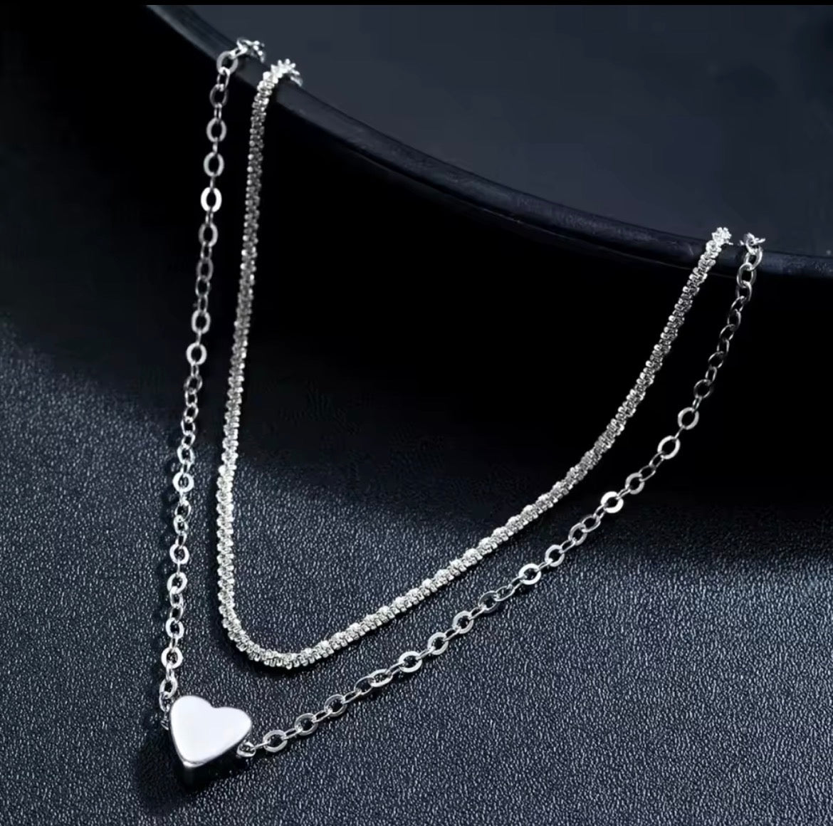 A silver double-layer necklace with a smooth heart pendant on a textured black background.