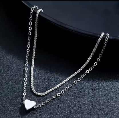 A silver double-layer necklace with a smooth heart pendant on a textured black background.