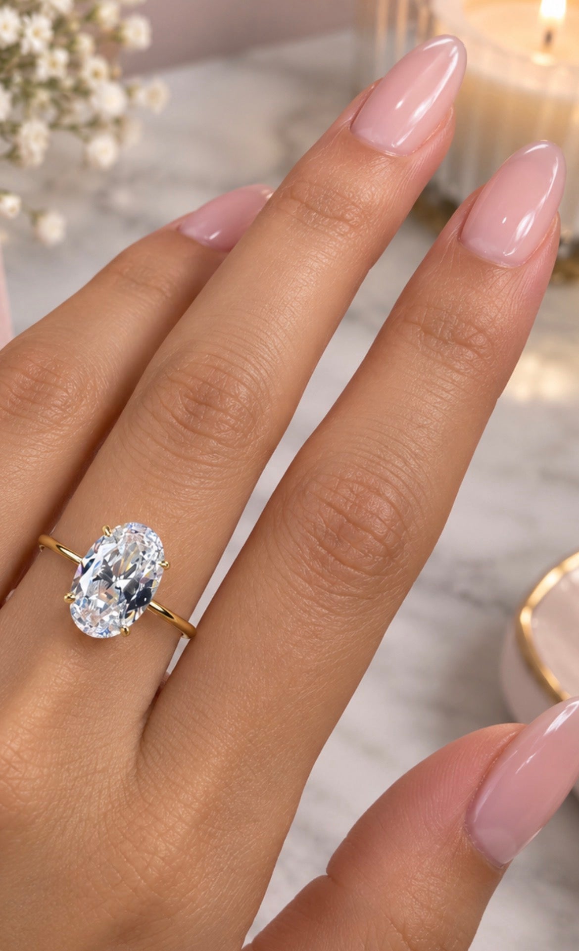A close-up of a hand with long, manicured pink nails wearing a gold ring with a large oval-cut clear gemstone, set against a softly blurred background with a candle and small white flowers.