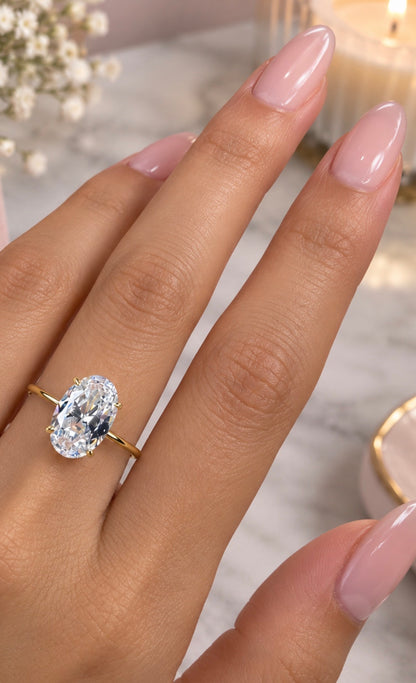 A close-up of a hand with long, manicured pink nails wearing a gold ring with a large oval-cut clear gemstone, set against a softly blurred background with a candle and small white flowers.