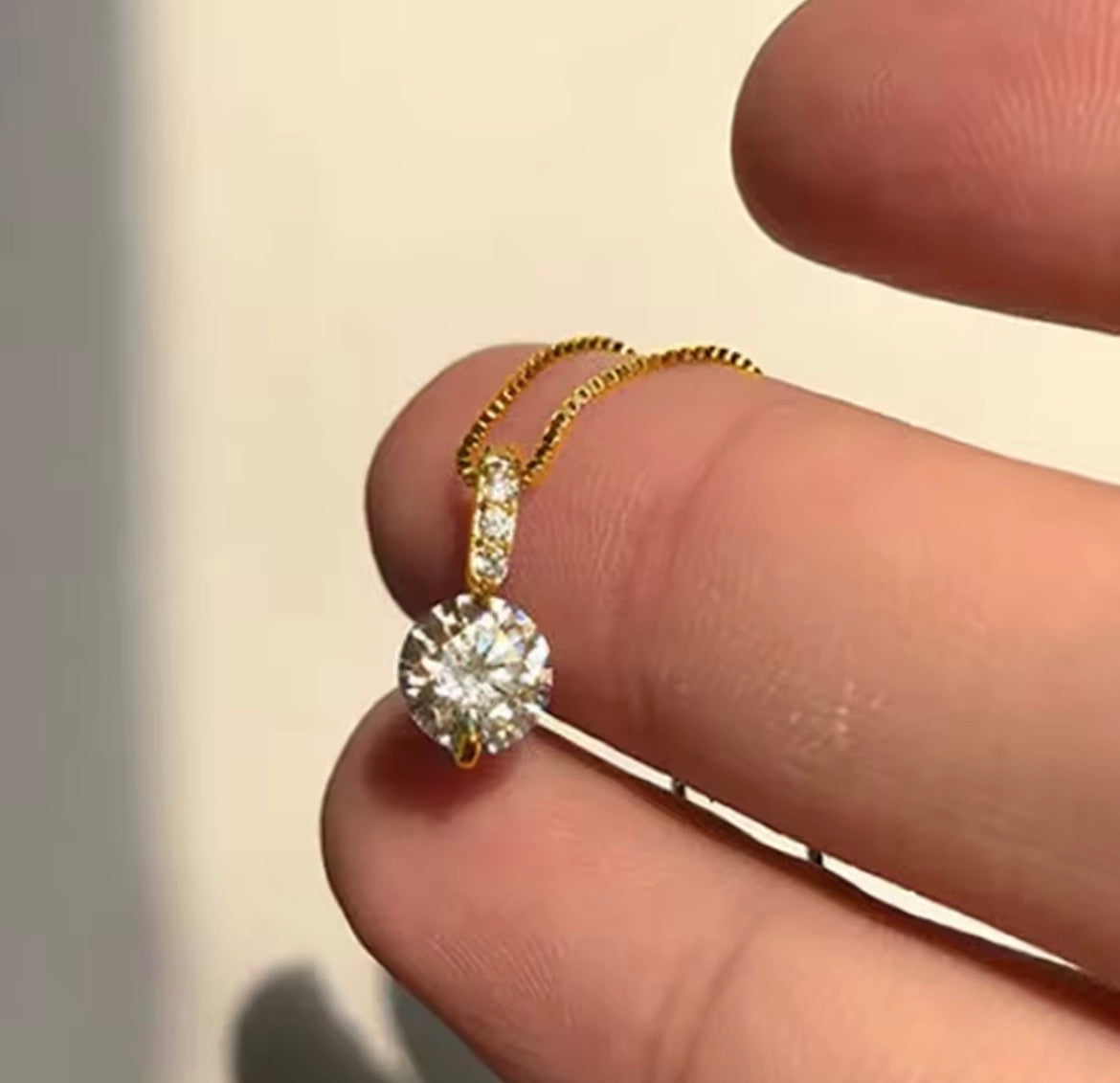 A close-up of a delicate gold necklace with a round-cut clear gemstone pendant, held between a person's fingers against a light neutral background.
