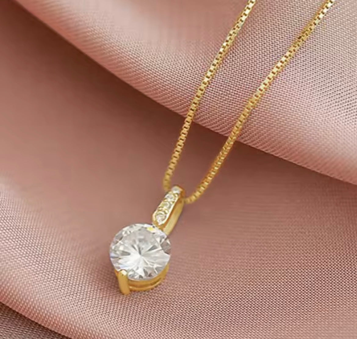 A gold chain necklace with a round clear gemstone pendant, displayed on draped pink satin fabric.
