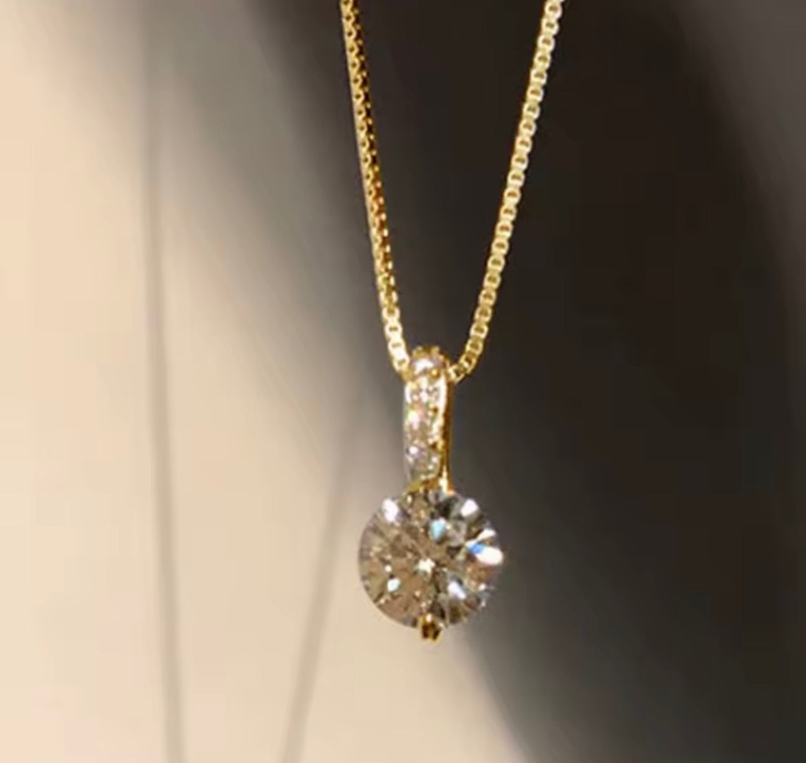 A gold necklace with a round-cut clear gemstone pendant, shown close-up against a dark and light gradient background.