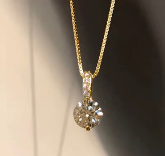 A gold necklace with a round-cut clear gemstone pendant, shown close-up against a dark and light gradient background.