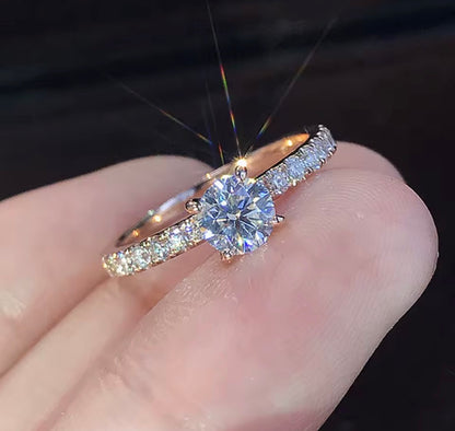A close-up of a rose gold ring with a round-cut center stone and pavé-set side stones, shown resting on a person's finger against a dark background.

