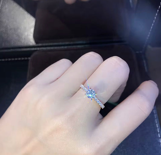A close-up of a hand wearing a delicate silver ring with a central clear gemstone and a pavé band, set against a dark background.
