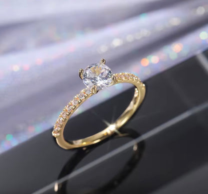 A gold solitaire engagement ring with a round clear center stone and a pavé-set band of small clear stones, displayed on a glossy black surface with a soft iridescent blurred background.