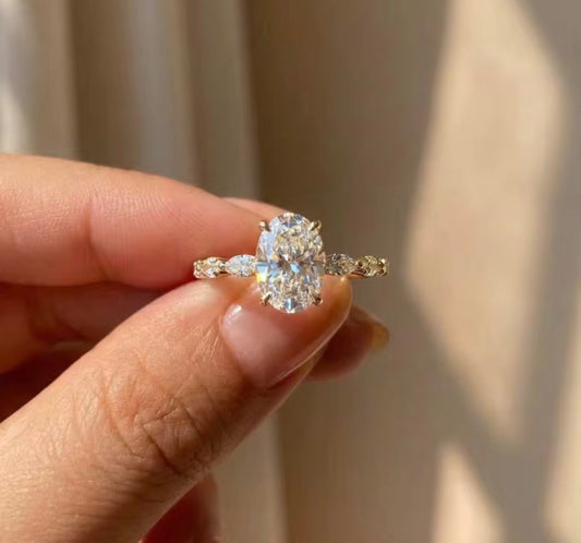 A close-up of a hand holding a gold engagement ring with a large oval center stone and smaller marquise-cut side stones, set against a soft beige curtain background.