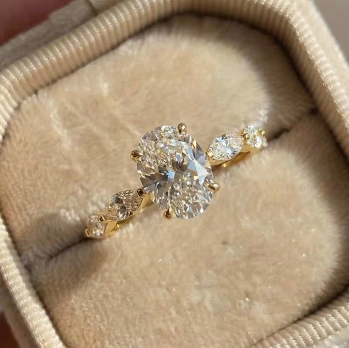 A yellow gold diamond ring with a large oval center stone and marquise-cut side stones, displayed in a beige velvet ring box.