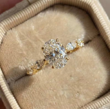 A yellow gold diamond ring with a large oval center stone and marquise-cut side stones, displayed in a beige velvet ring box.