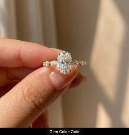 A close-up of a hand holding a gold engagement ring with an oval center stone and marquise side stones, with a text overlay at the bottom reading 'Gem Color: Gold'.