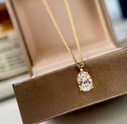 A gold chain necklace with an oval-cut clear gemstone pendant, resting on a brown jewelry box.
