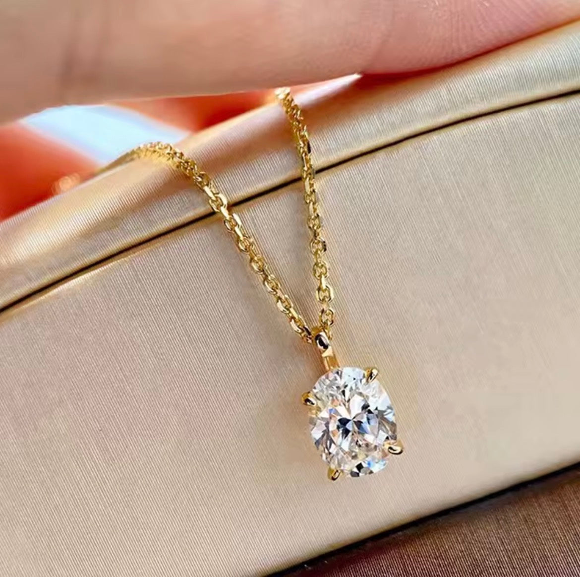 A gold solitaire necklace with a round-cut clear gemstone pendant, resting on a beige satin jewelry box with a hand holding the chain above.