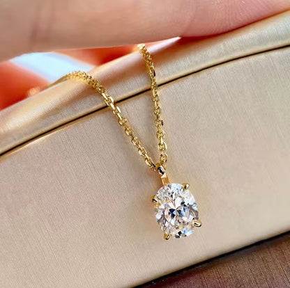 A gold solitaire necklace with a round-cut clear gemstone pendant, resting on a beige satin jewelry box with a hand holding the chain above.
