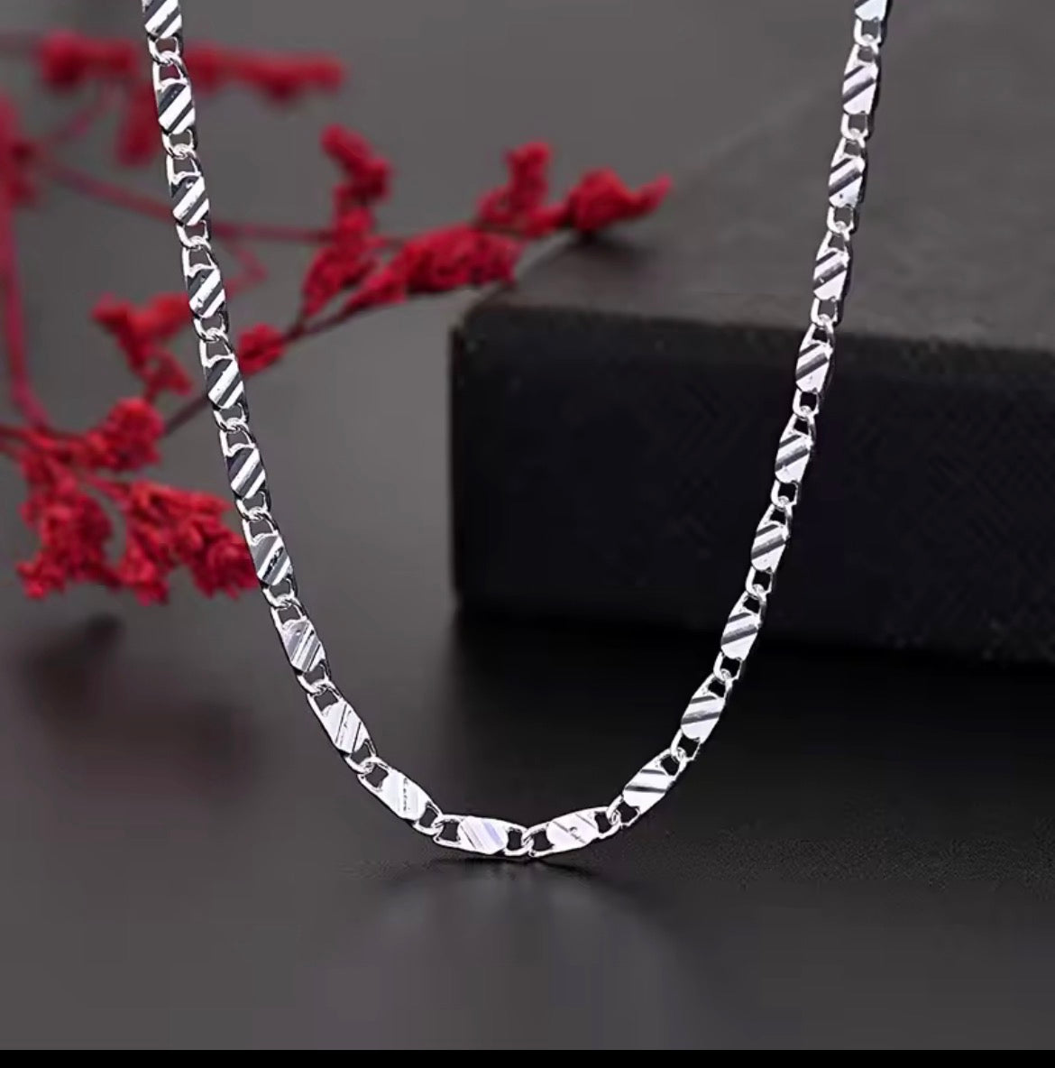 A silver chain necklace with a textured, diamond-cut link design, draped elegantly over a dark surface with a black box and red decorative flowers in the background.
