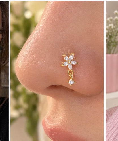 Close-up of a gold-tone nose stud worn on a nostril, featuring a five-petal clear-stone flower and a dangling round clear stone.