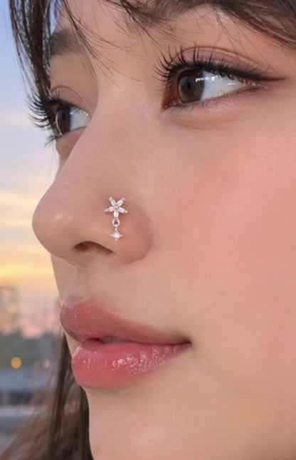 A close-up of a woman's face wearing a delicate silver nose stud with a small flower and dangling charm, set against a softly blurred outdoor background at sunset.