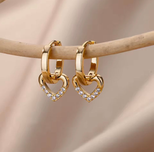 A pair of gold huggie hoop earrings with interlocking heart charms set with small clear stones, hanging from a light wooden branch against a soft beige fabric background.