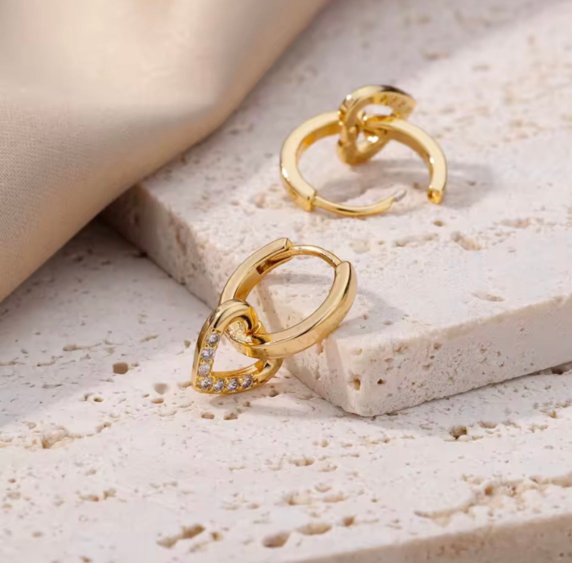 A pair of gold huggie hoop earrings, one adorned with a small heart-shaped charm set with clear crystals, displayed on a textured beige stone block with a draped beige fabric.