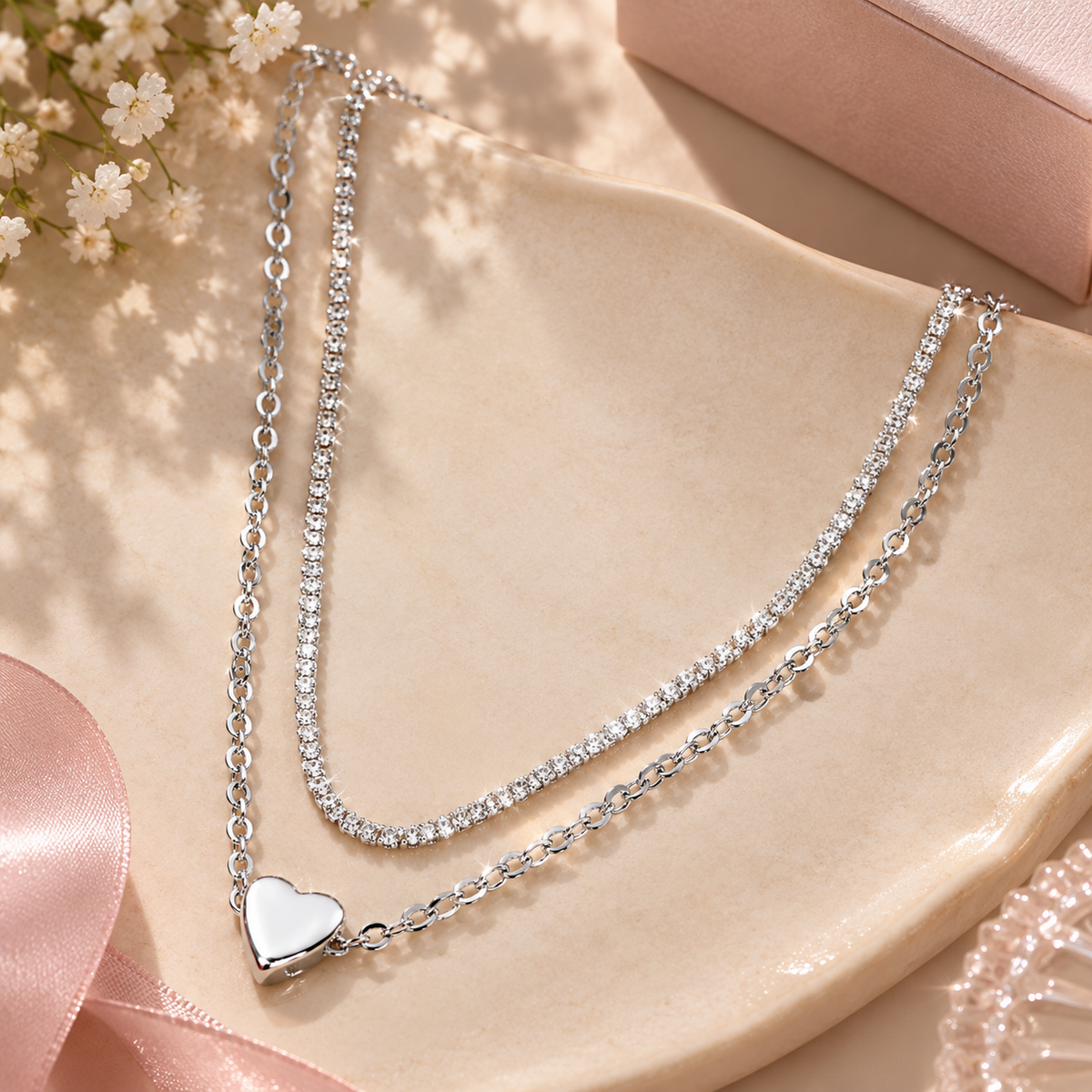 A double-strand silver necklace with clear rhinestone-studded chains and a polished silver heart pendant, styled on a beige ceramic dish with pink ribbon and small white flowers.