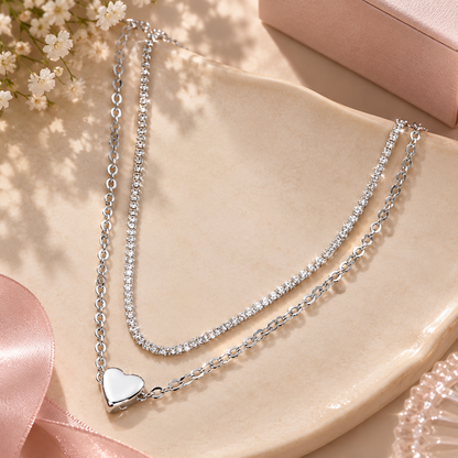 A double-strand silver necklace with clear rhinestone-studded chains and a polished silver heart pendant, styled on a beige ceramic dish with pink ribbon and small white flowers.